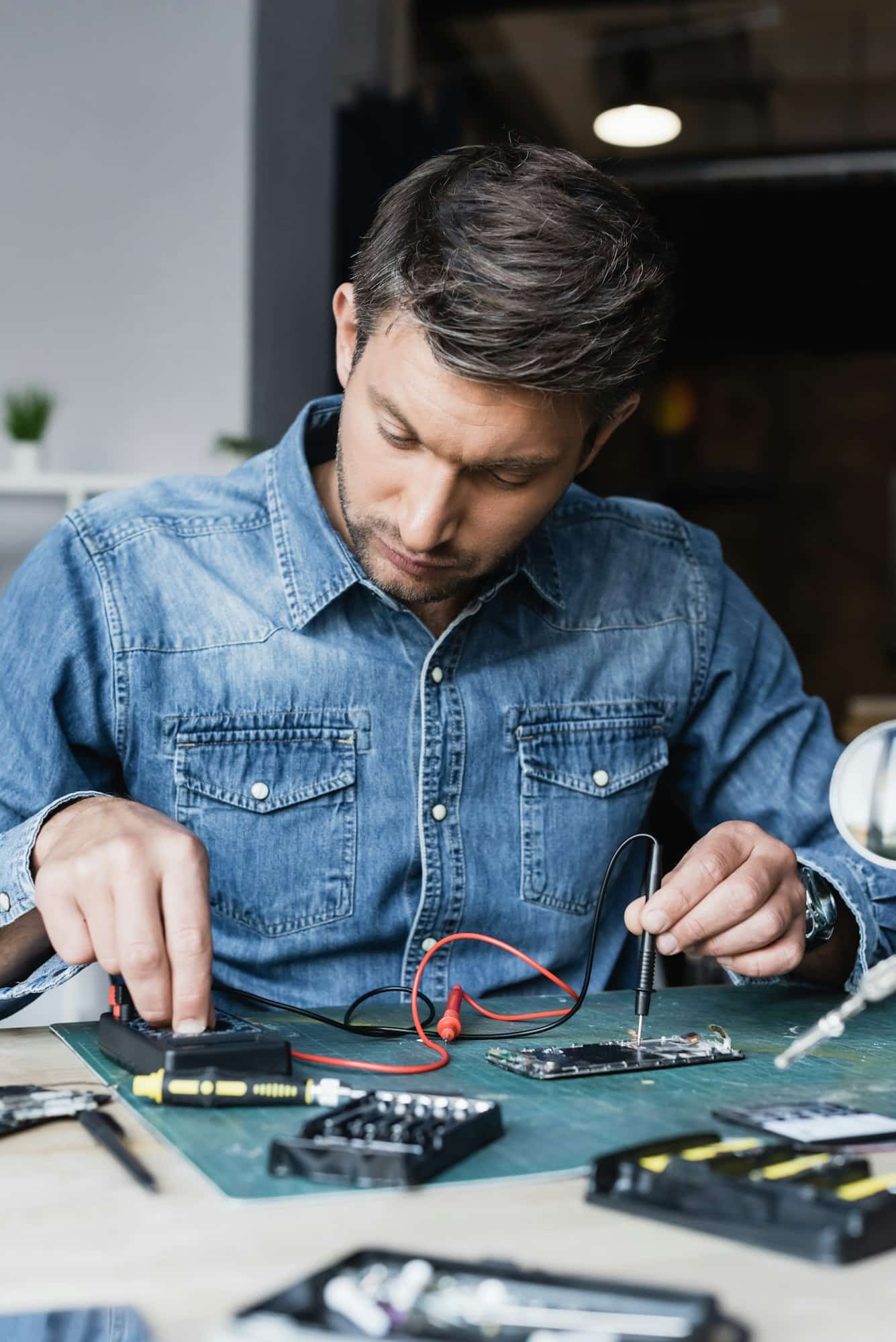 Focused repairman regulating multimeter while holding sensor on disassembled part of mobile phone on